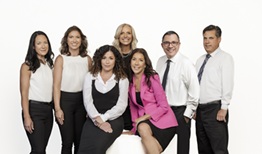 A diverse group of seven professionals posing together in front of a white background. They are dressed in a mix of business attire, including a pink blazer. The group includes men and women with varying hair styles and colors.